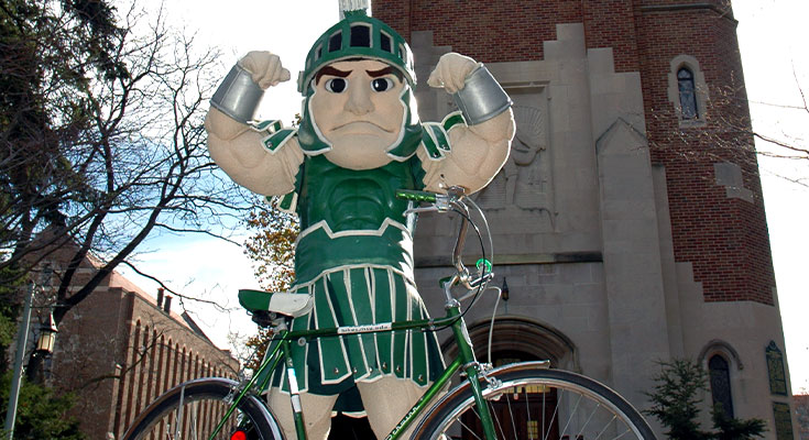 A photo of Sparty posing infront of Beaumont Tower with his bicycle.