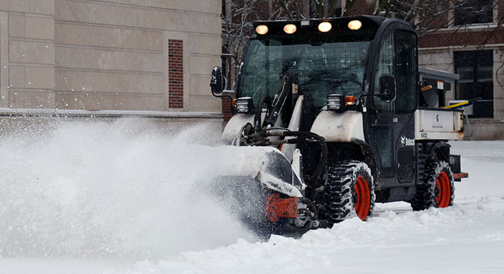 A photo of a Landscape Services employee operating snow broom equipment on a campus sidewalk.