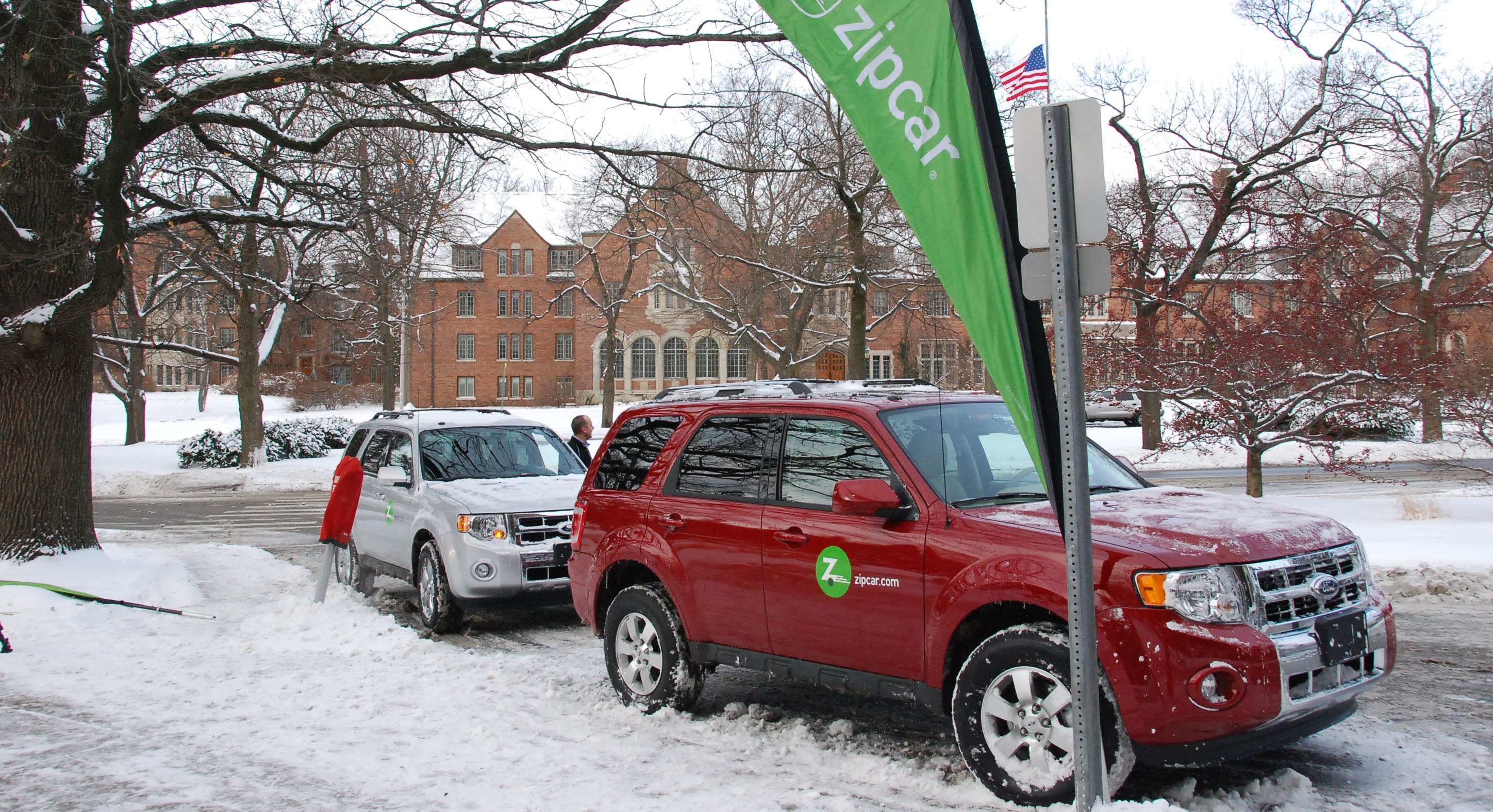 One white Ford Escape and one red Ford Escape are parked in front of the MSU Union by a Zipcar sign. Campus is snowy.