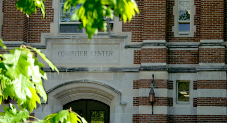 A photo of the Computer Center main entrance behind beautiful green leaves on a branch outside.