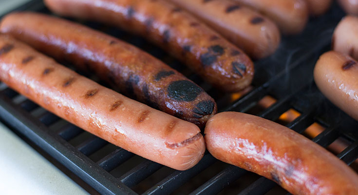A photo of hot dogs cooking on a portable propane grill. 